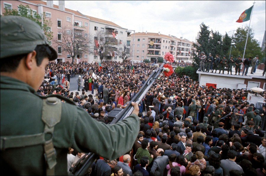 A soldier holds a rifle with several carnations attached to it, standing above a large crowd in a city square.