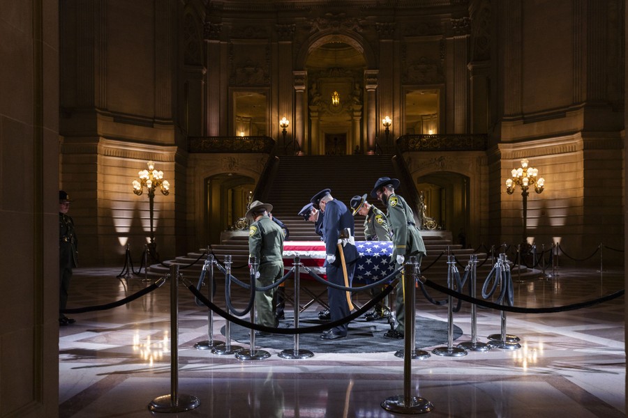 Six uniformed people guide a casket draped with an American flag inside a city hall.