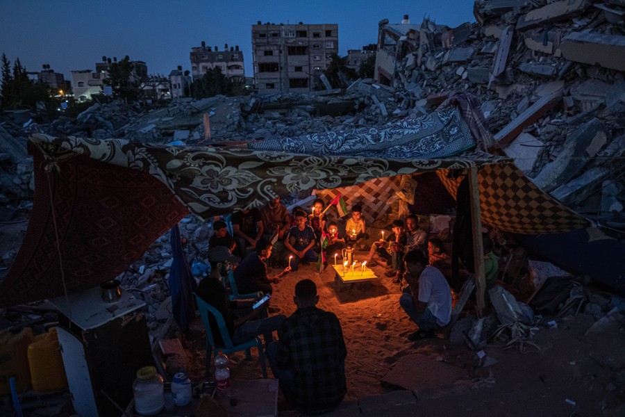 About a dozen children are seen sitting and holding candles inside a makeshift tent among piles of rubble.
