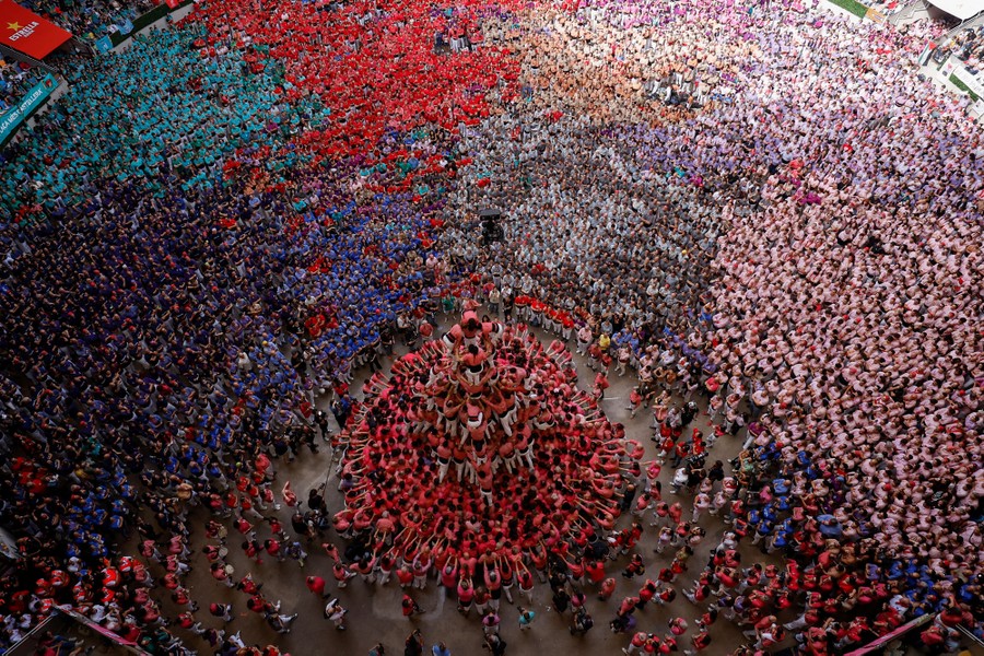 A large arena space is filled with about eight or nine teams of people, all watching another team form a human tower.