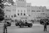 This black and white photo shows men in combat attire standing guard outside a large brick school building. Three military-esque Jeeps are in the photo.