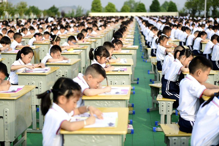 Hundreds of young students sit at desks lined up outside.