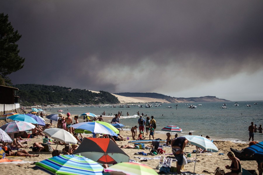 Many people play and relax on an ocean beach as a black cloud of smoke rises in the background.