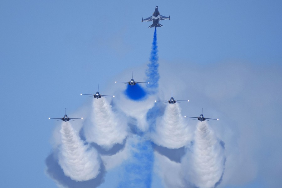 Six military jets fly in formation, releasing colored smoke behind them.