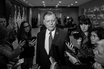 Black-and-white photograph of Tom Cole, dressed in a suit, surrounded by reporters holding their phones up toward him.