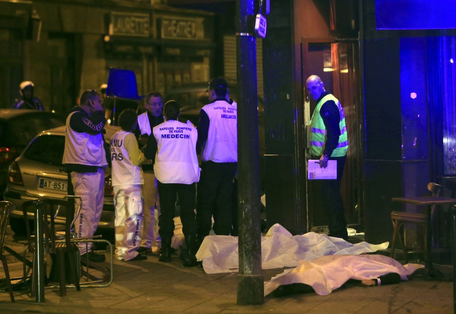 Medical personnel stand by two bodies covered in white sheets in a city street.