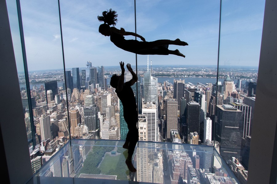 A person tosses another person above them, while standing in an all-glass observation deck high above New York City.
