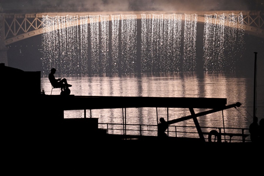 A man sits in a chair atop a boat against the backdrop of fireworks cascading from a nearby bridge.