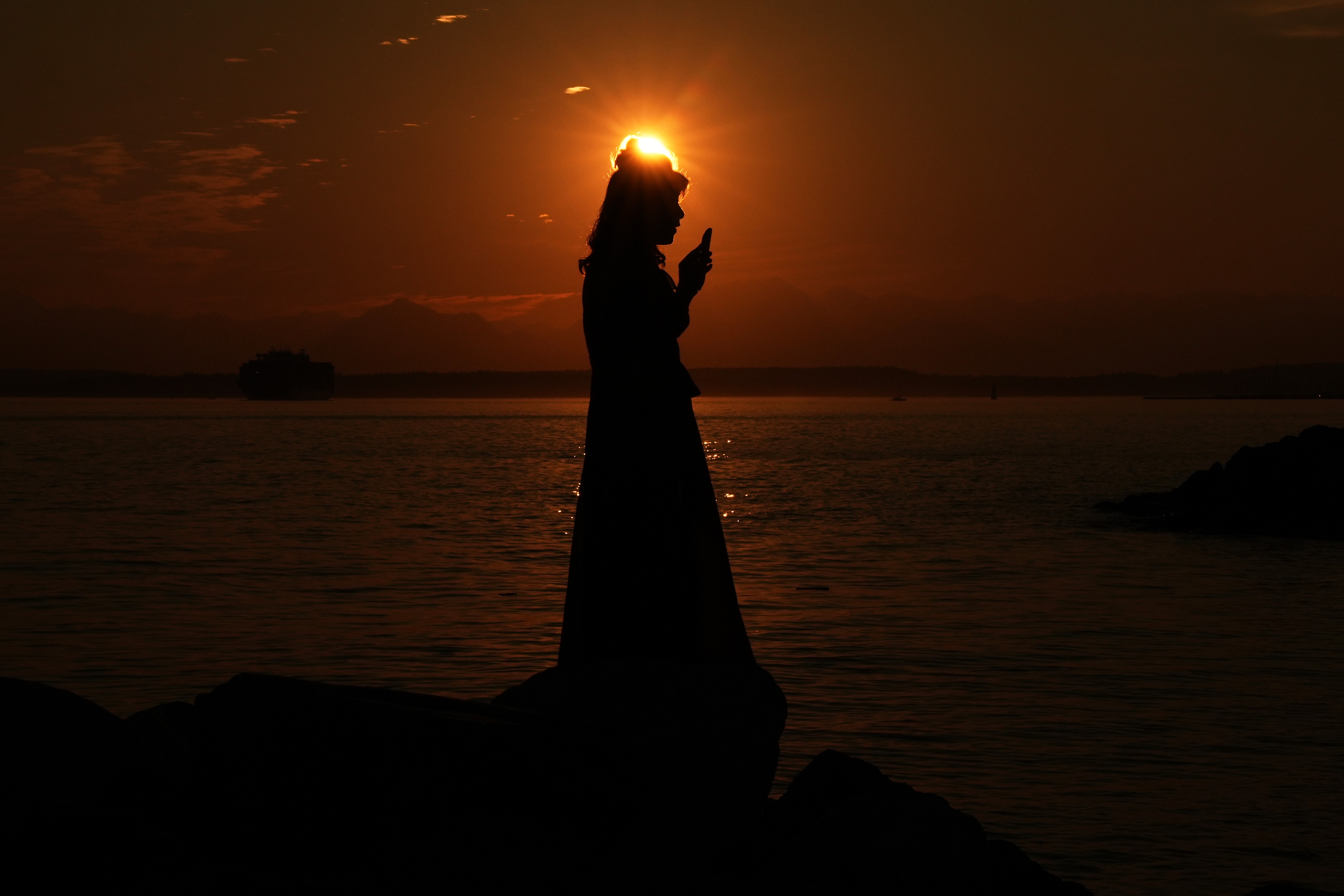 A woman is silhouetted against the setting sun while holding up her phone.