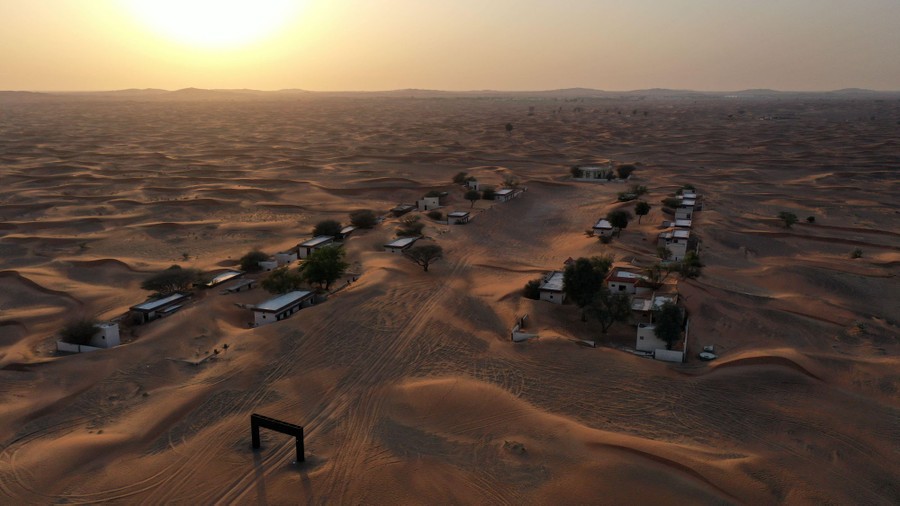 A couple of dozen small buildings stand clustered together in a desert, surrounded and half-buried by sand dunes.