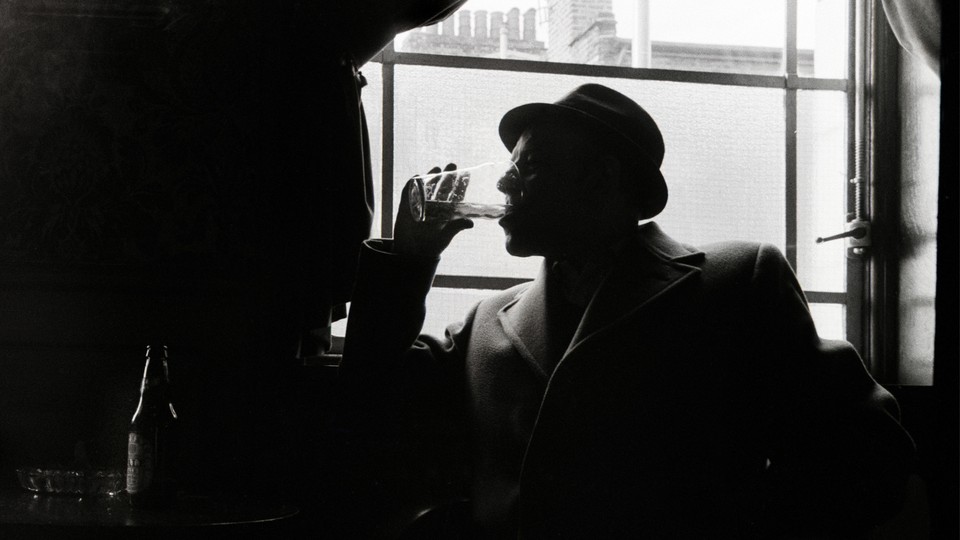 A man wearing a hat and a wool coat drinks from a glass while he sits next to a window