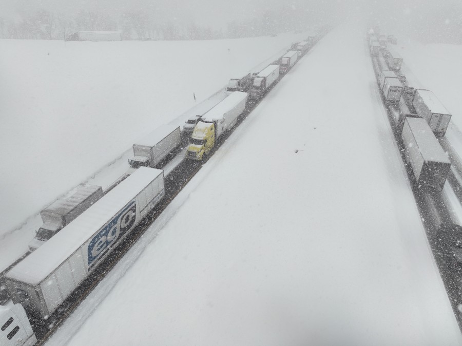 An aerial view of a snow-covered highway, filled with stopped trucks in both directions