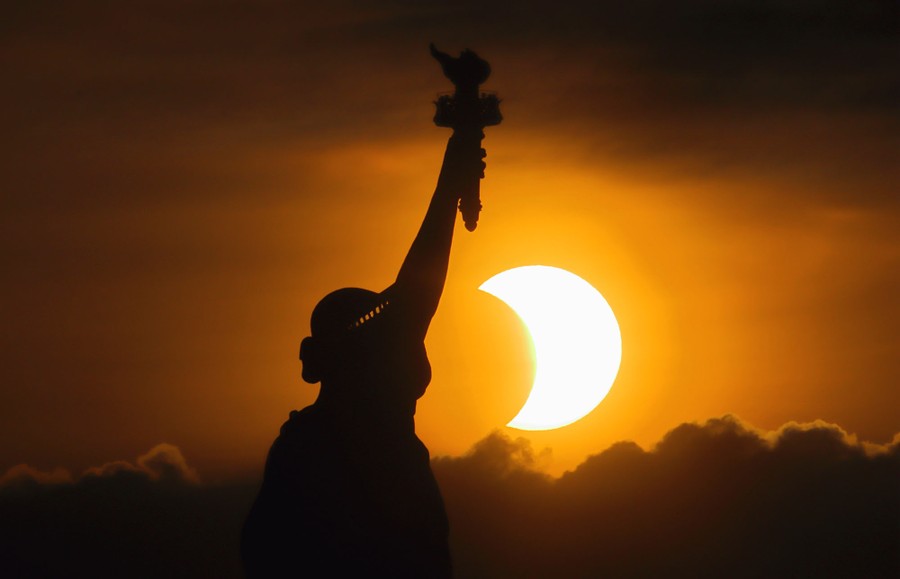 A partly-eclipsed sun, seen behind the Statue of Liberty.