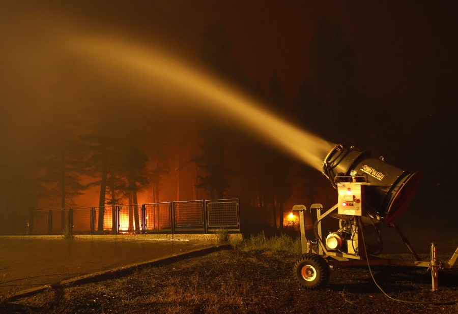A snow-making machine sprays water into the air near a burning forest fire.