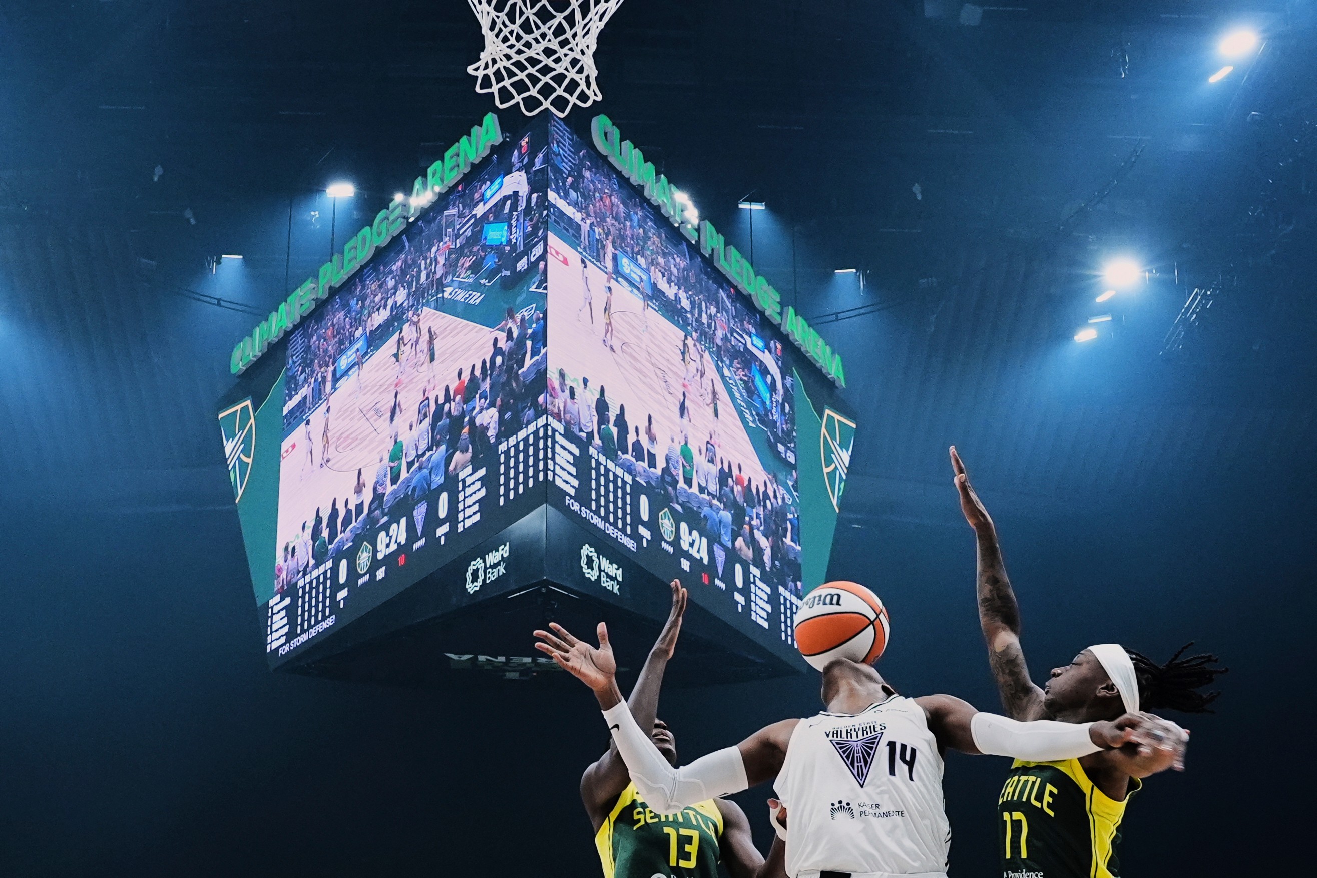 Three basketball players vie for a loose ball, seen beneath a large central video display installation.