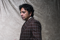 portrait of M. Night Shyamalan standing, looking down with hands clasped, wearing brown plaid suit