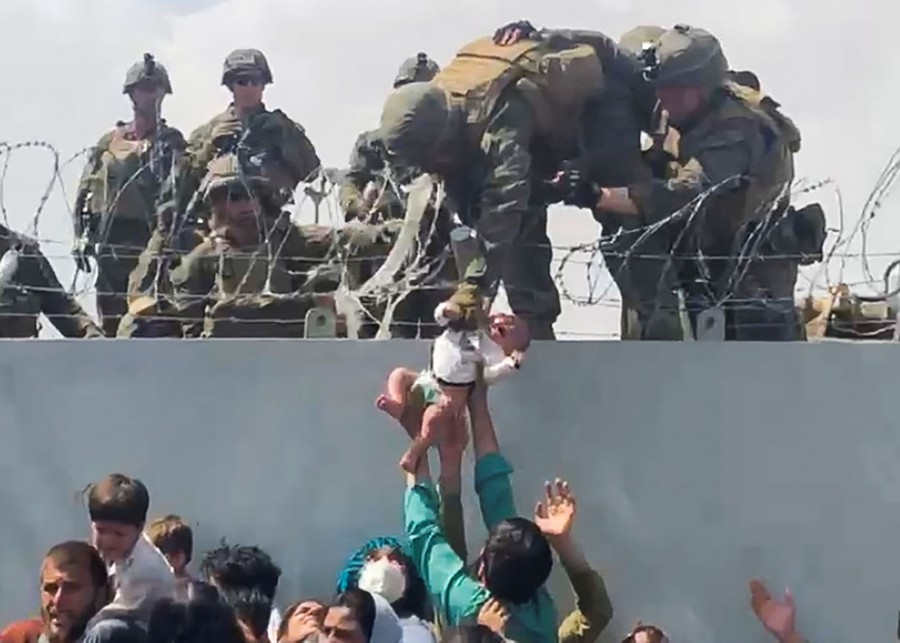People reach up to pass a baby to soldiers standing atop a wall edged with razor wire.