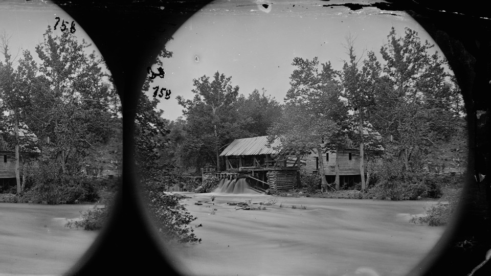 A black-and-white photograph of a house with a stream running near it, surrounded by trees and water