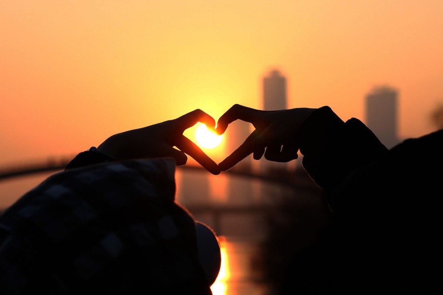 People make a heart shape with their fingers to capture the sun rising in the background.