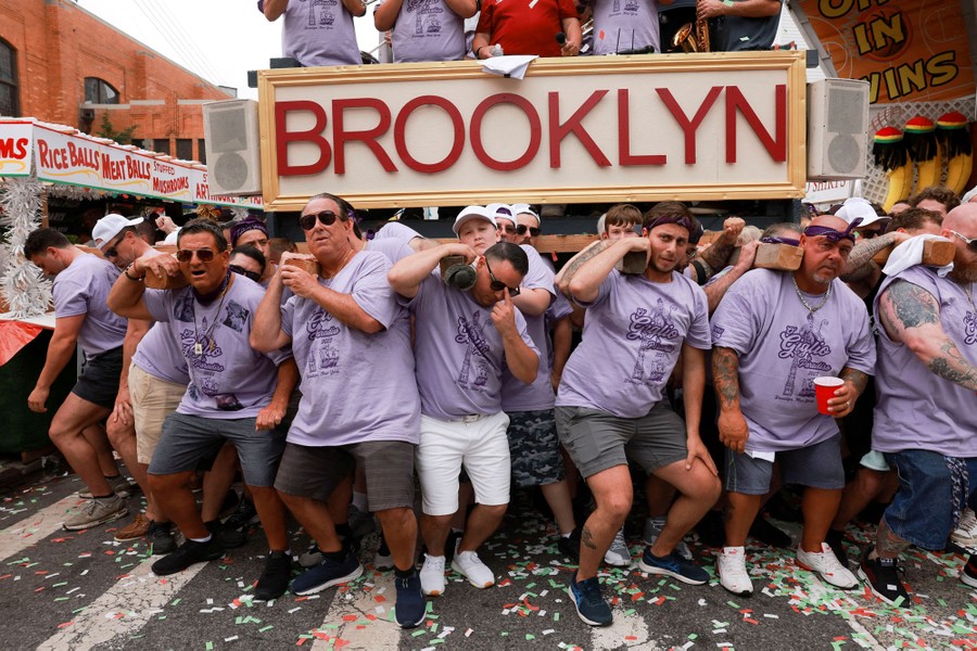 People crouch while using their shoulders to lift a large float with the word "Brooklyn" written across the front.