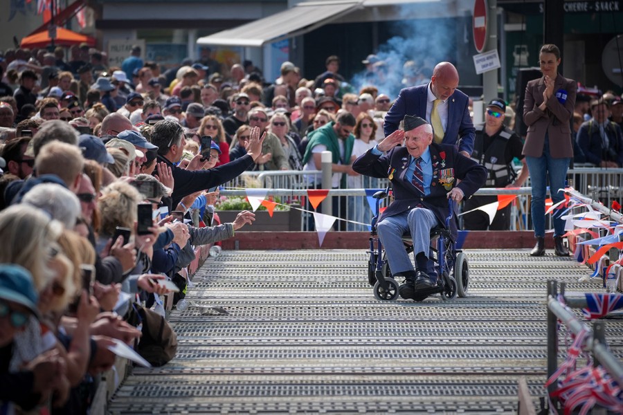Crowds applaud an older veteran as he is pushed past them in a wheelchair.