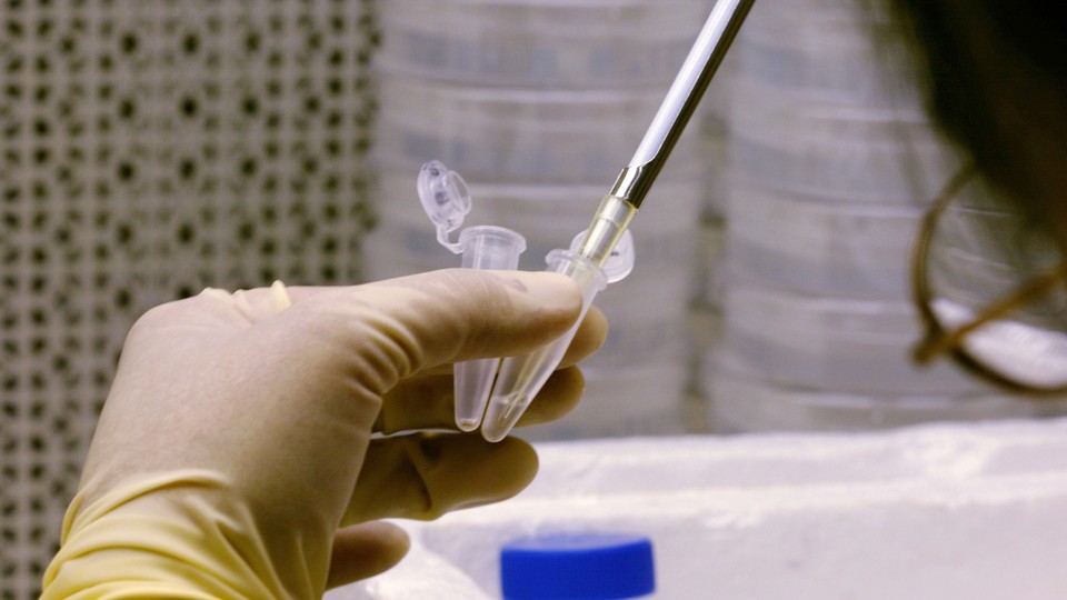 A person holds two vials in a gloved hand while inserting a pipette into one vial.