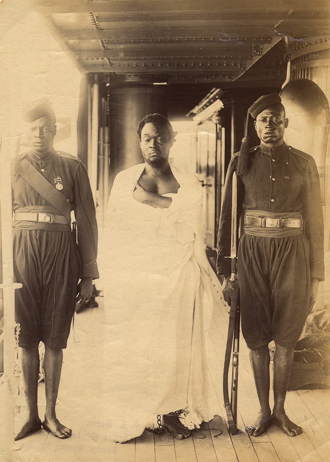 sepia-toned archival photo of a man in white robes and ankle shackle standing between two men standing at attention with rifles