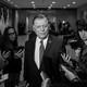 Black-and-white photograph of Tom Cole, dressed in a suit, surrounded by reporters holding their phones up toward him.