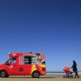 A woman pushing a red pram toward a red ice-cream truck on a beach.