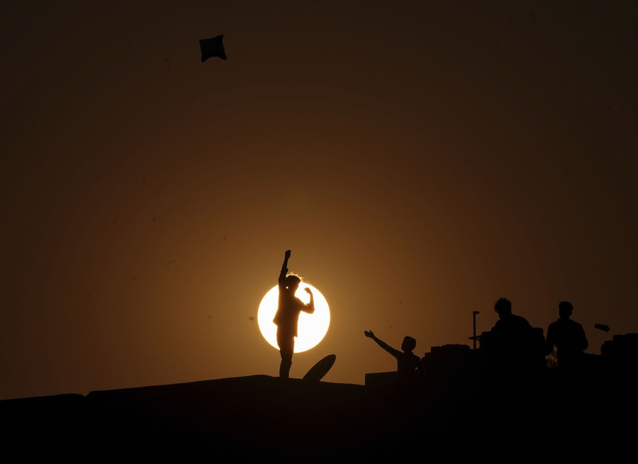 People on a rooftop are silhouetted by the sun while flying kites.