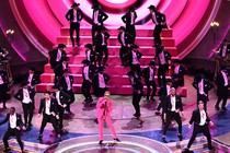 Ryan Gosling, dressed as Ken in a pink suit and gloves, sings into a microphone on the Oscars stage as several dozen men in black tuxedos dance behind him.