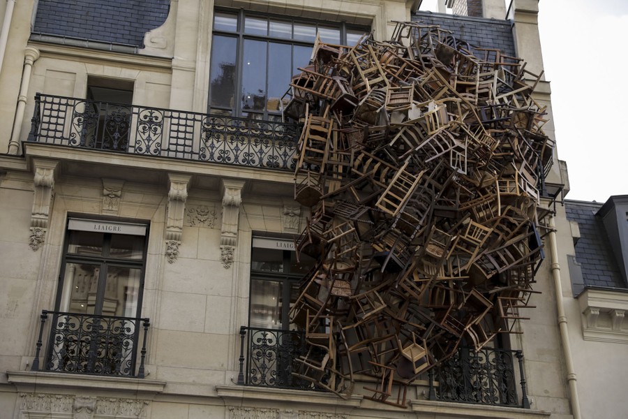 Dozens of wooden chairs are clustered into a tangled bunch, attached to the side of a mansion in Paris.
