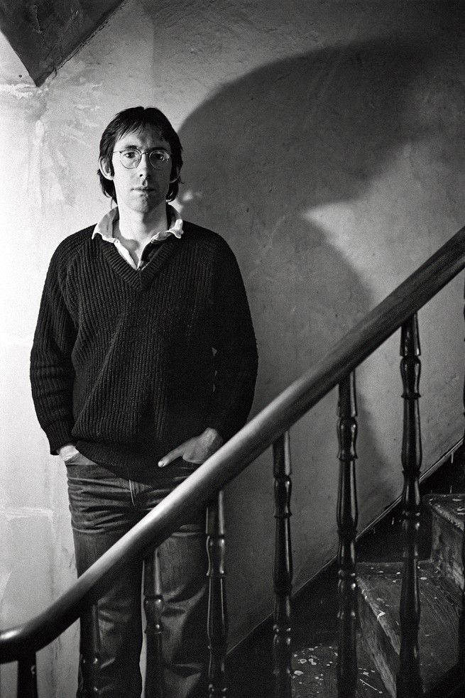 black and white photo of man wearing glasses, heavy sweater, and jeans with hands in pockets, standing on steps in shadowy stairwell behind a bannister