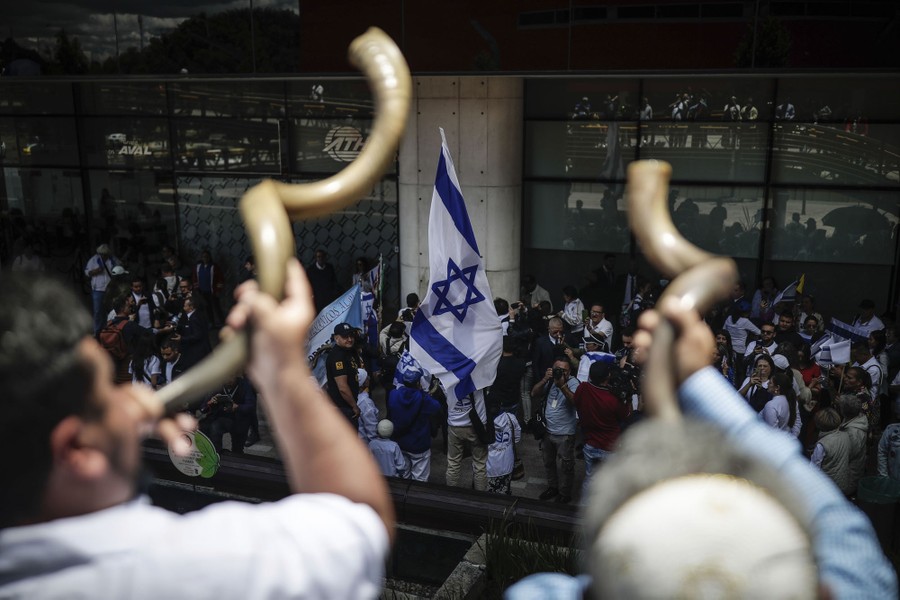 Two people blow shofars as a crowd carrying Israeli flags gathers outside a building.
