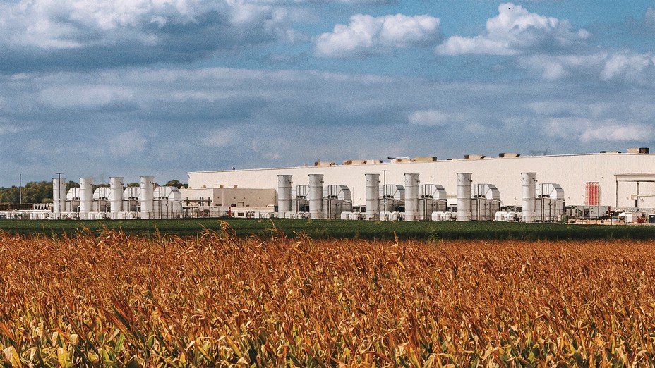 photo of enormous warehouse with numerous external cooling structures, with bronzed field of corn growing in foreground