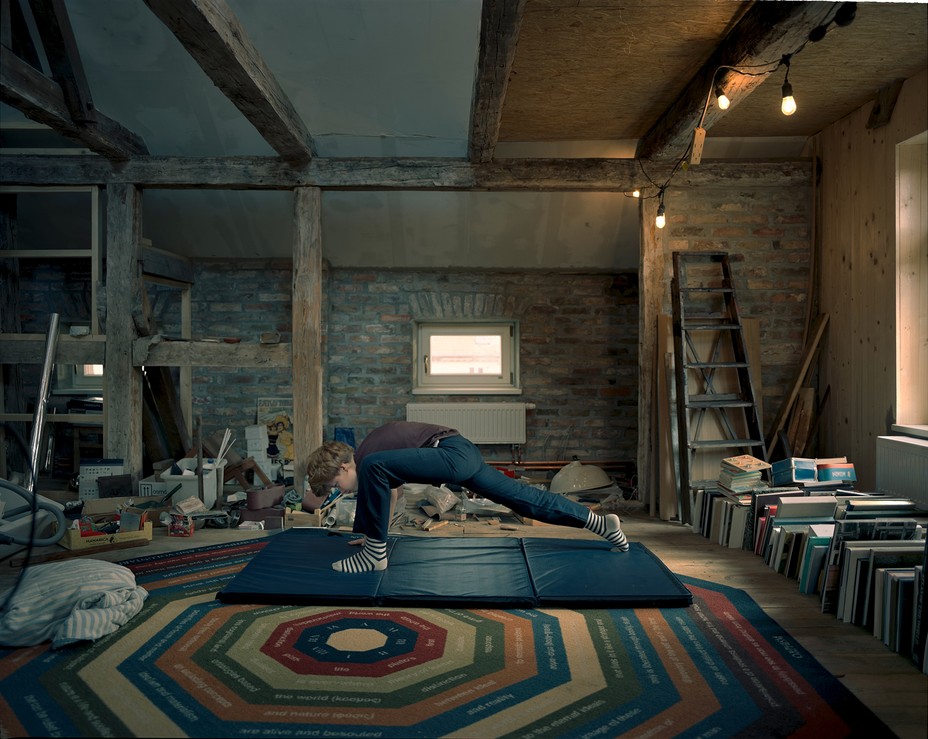a boy stretches in a lunge on a gym mat surrounded by books in wood-beamed room of stone house