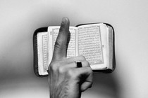 A photograph of a raised finger in front of a Quran