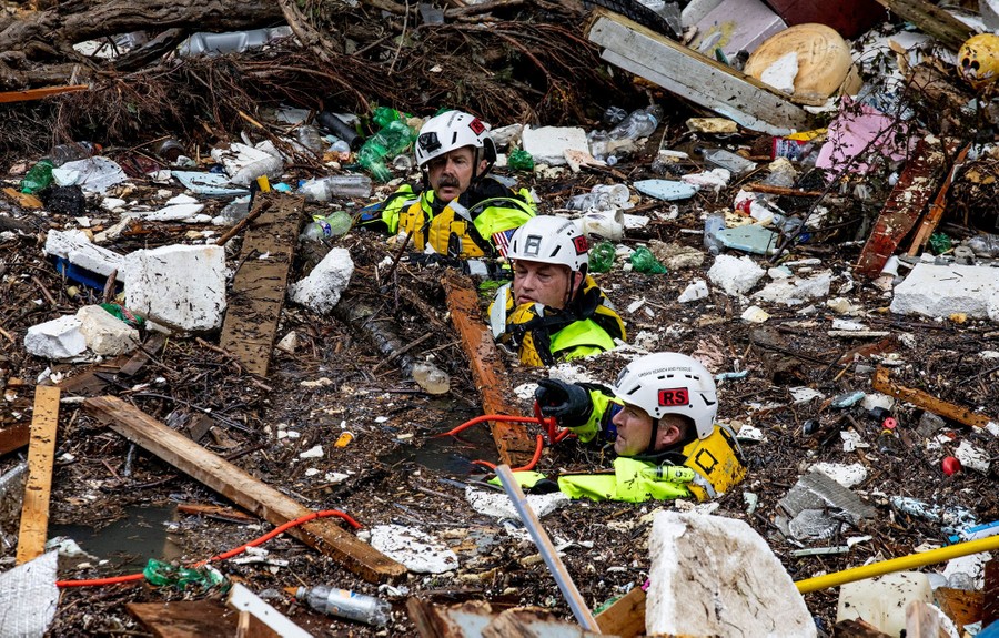 Three rescue workers struggle in deep water, surrounded by flood debris.