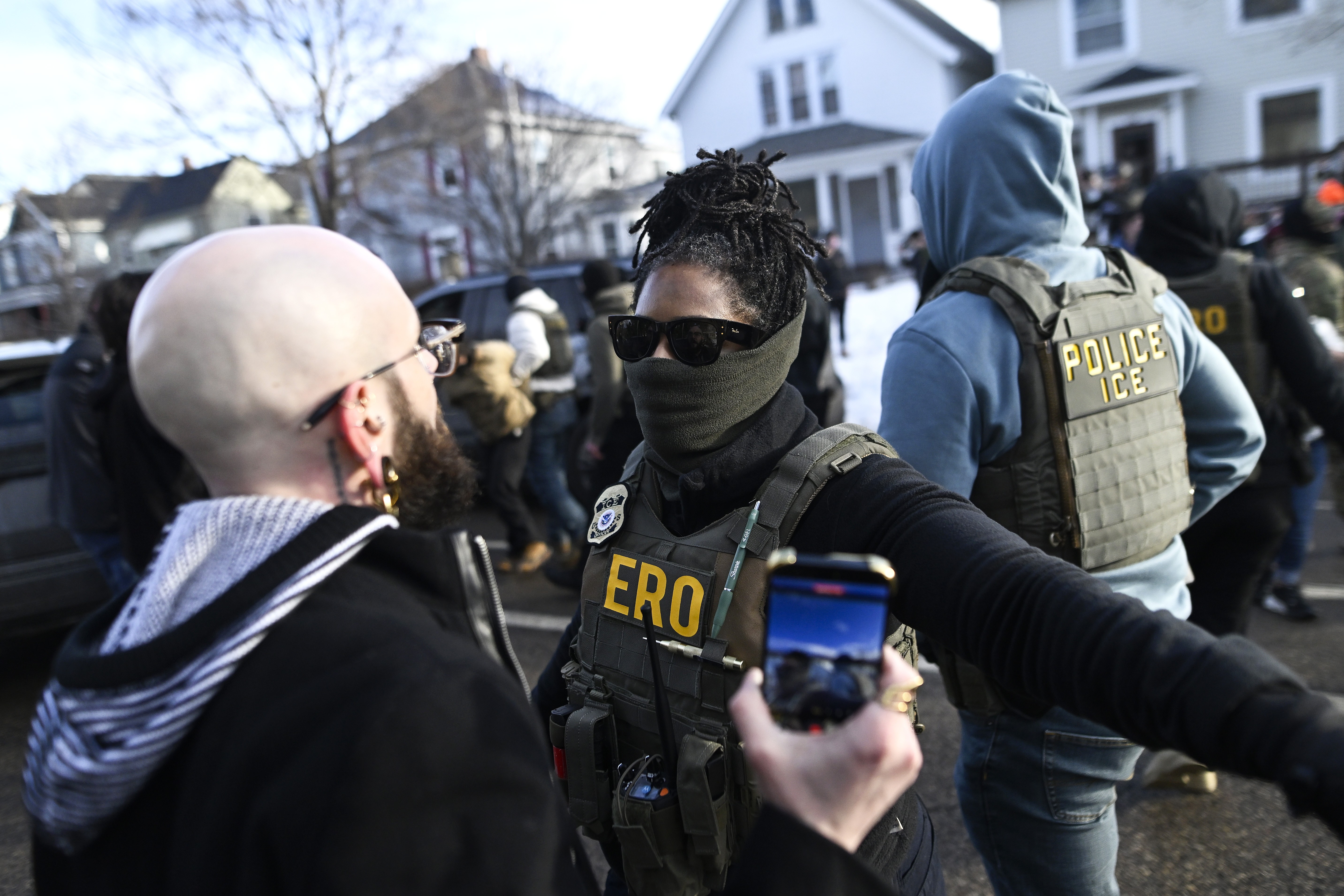 A federal agent wearing a mask holds out their arm, trying to hold back civilians who gathered at the scene of an immigration arrest.