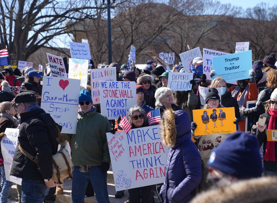 Protesters gather, holding signs, including one that reads 