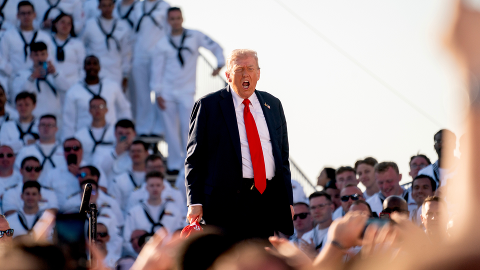 President Donald Trump is shown speaking, with his mouth open, surrounded by uniformed members of the U.S. Navy.