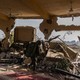 A soldier walks through a blown out interior in Gaza, ceiling fans and carpets visible under rubble but walls blasted through.