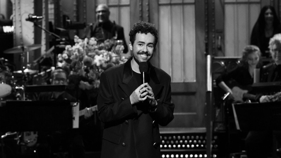 Ramy Youssef smiles on the SNL stage while holding a microphone