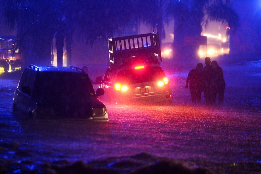 Several people walk near two stranded cars during a flash flood at night.