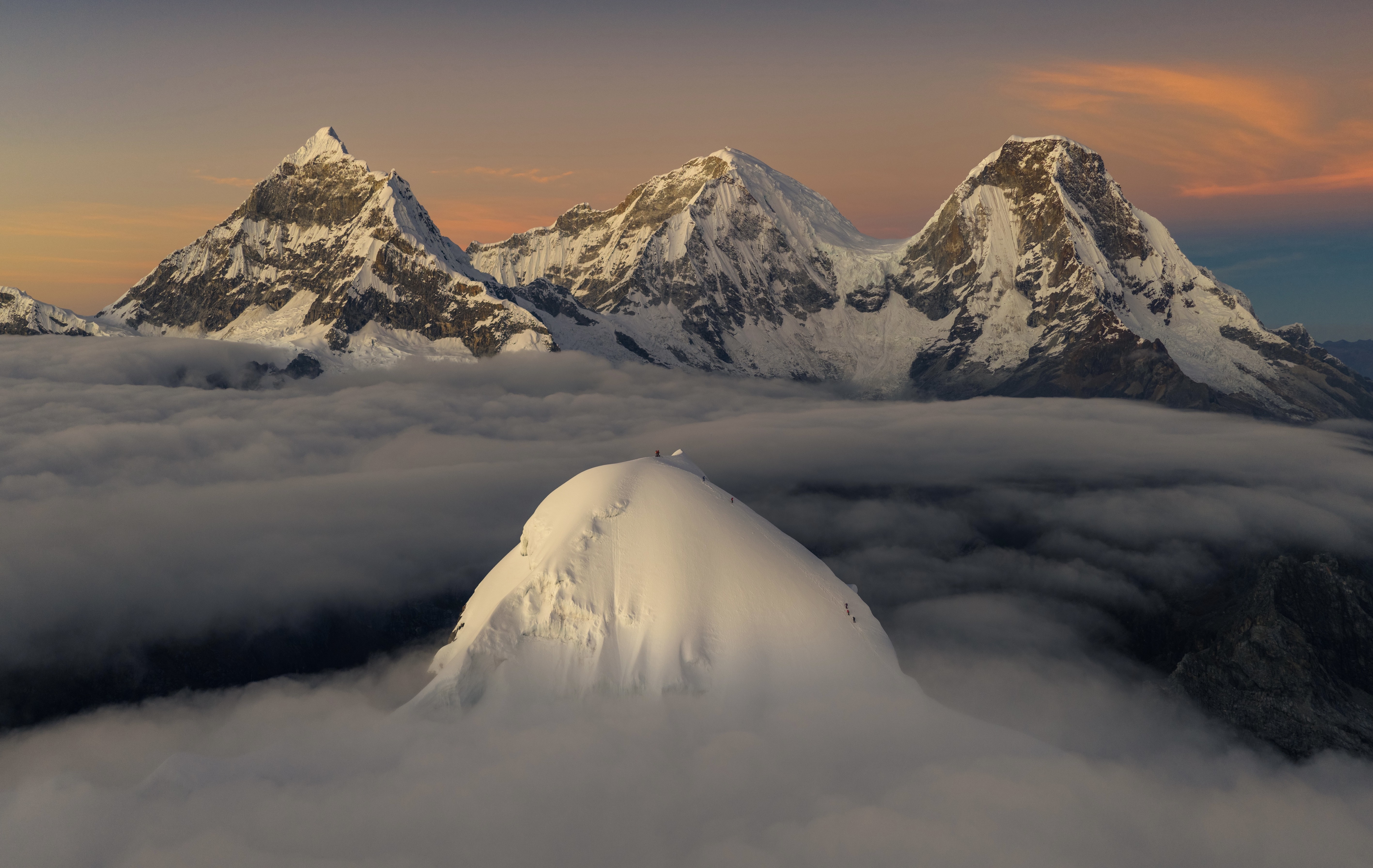 A half-dozen mountain climbers, seen as tiny specks, reach the summit of a snow-covered peak, with much-taller jagged mountains in the background.