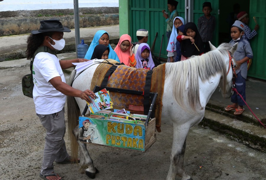 A person reaches for books that have been carried to a village in boxes carried by a horse, while about a dozen small children wait.