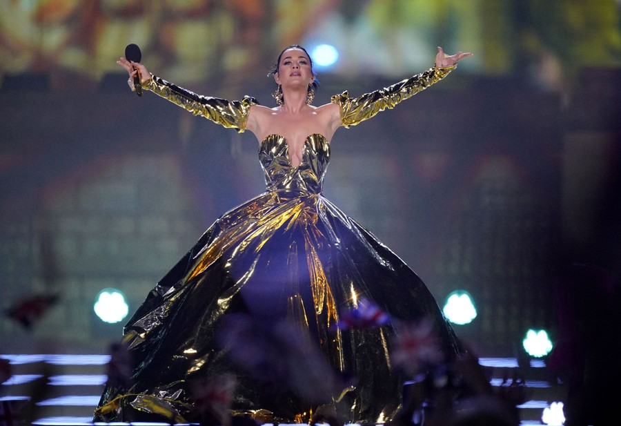 Katy Perry stands onstage in a full, shiny dress during a performance, arms held wide.