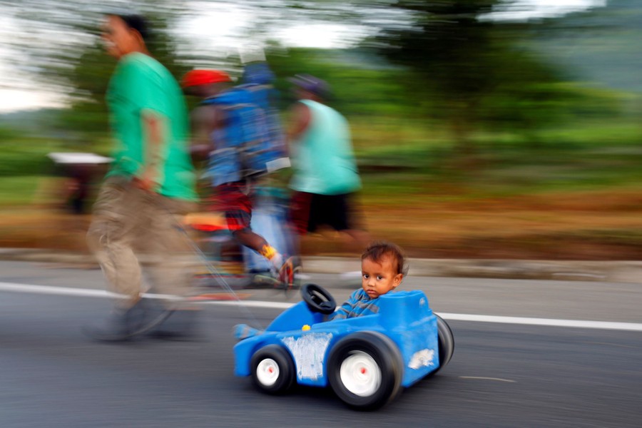 A young boy rolls down a road in a toy car pulled by his father.