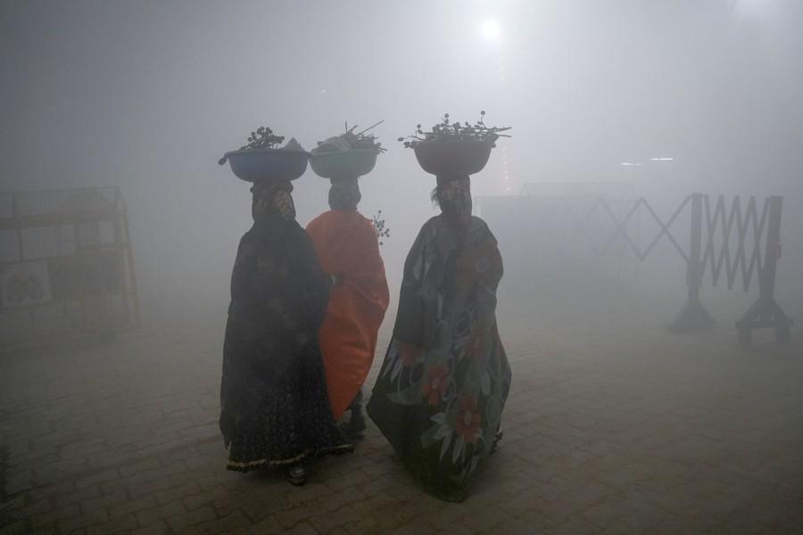 Three people carrying tubs full of branches on their heads walk through dense fog.