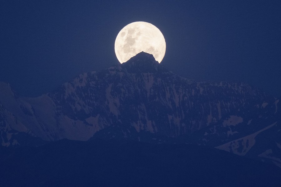 A full moon rises over the peak of a tall mountain.
